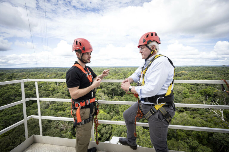 The President of Germany listens to scientist Stefan Wolff, who explains the ATTO research. Both stand on the 54 m platform of the ATTO tower, wearing bright red helmets and safety belts.