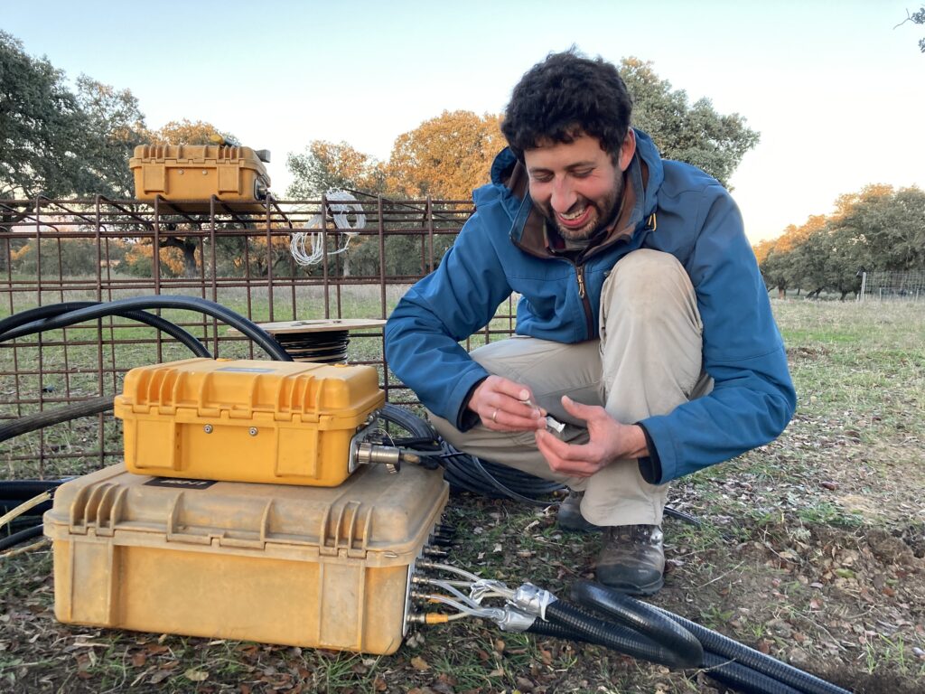 Tarek crouches on a meadow next to a box of instruments and checks the connections.