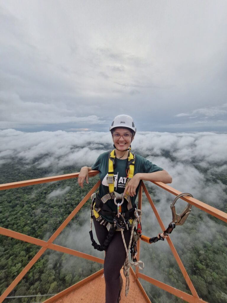 A woman is standing on a platform, forest and clouds are below her. She's wearing a hard hat and is secured to the platform with a harness.