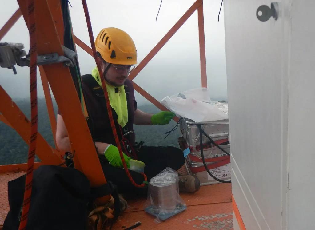 Man in safety gear working on a tall tower with equipment.
