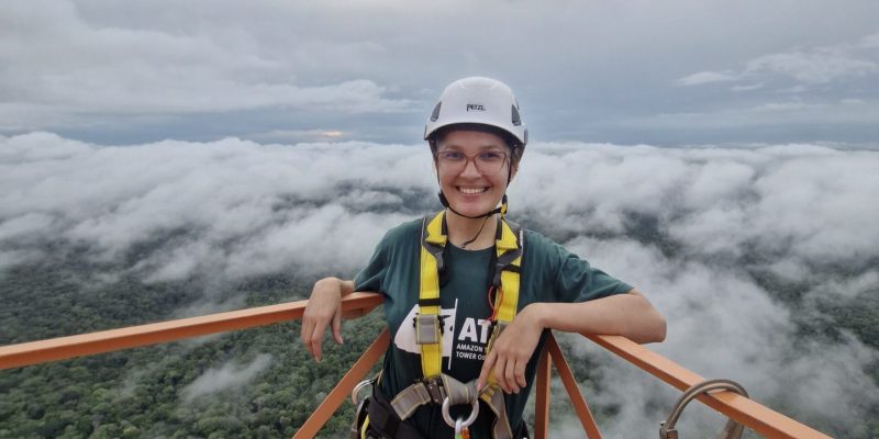 A woman is standing on a platform, forest and clouds are below her. She's wearing a hard hat and is secured to the platform with a harness.