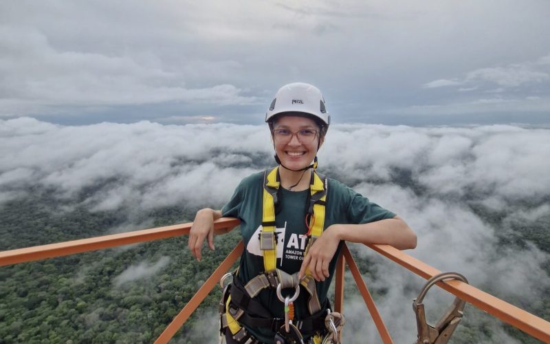 A woman is standing on a platform, forest and clouds are below her. She's wearing a hard hat and is secured to the platform with a harness.